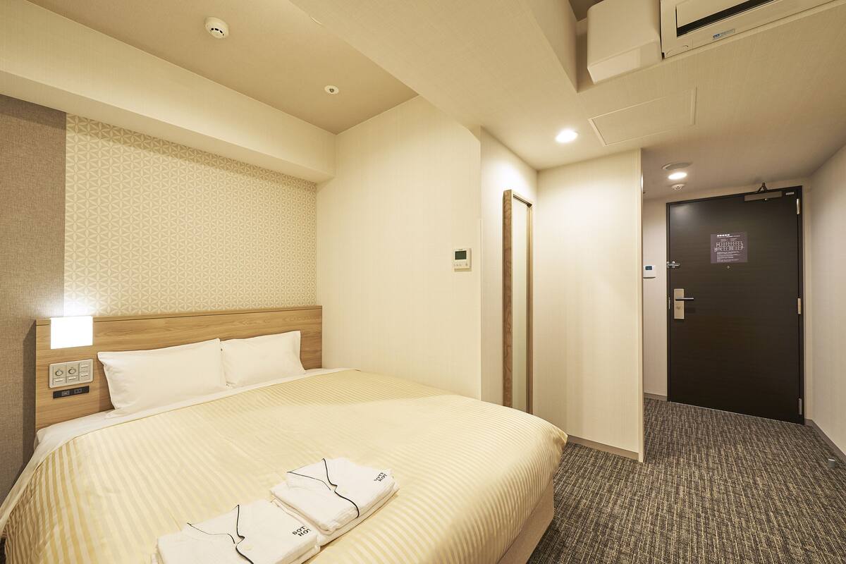 A hotel room with a large bed, a bedside table with a lamp, a wall-mounted control panel, and a door with a sign.