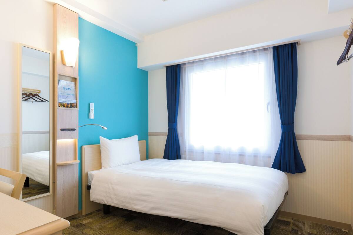 A hotel room with a bed, a desk, a mirror, a wardrobe, and a window with curtains.