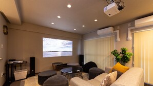 En moderne stue med sofa, skottomaner, et sofabord og et projektorskærm, der viser et naturskønt udsigt.