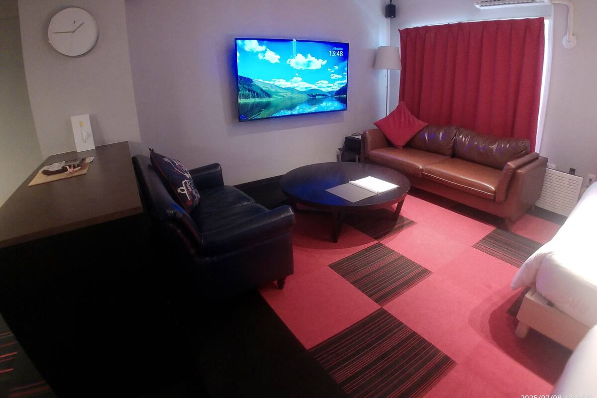 A hotel room with a sofa, a coffee table, a TV mounted on the wall, and a red curtain.