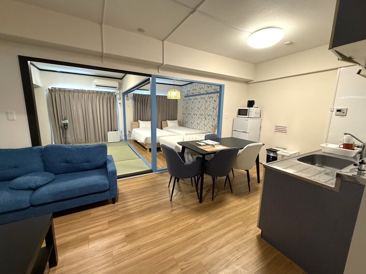 A compact hotel room with a sofa, a dining table with chairs, a kitchenette, and a separate bedroom area with a bed and curtains.