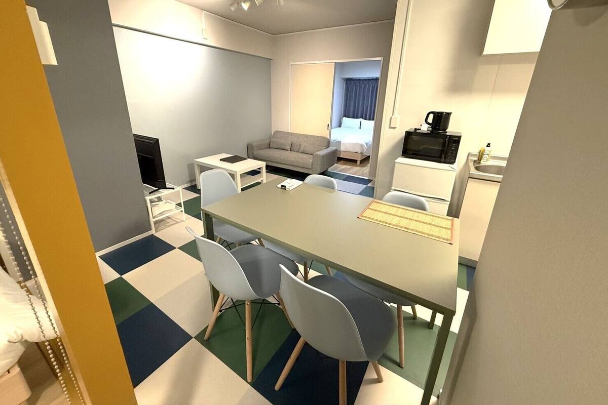 A compact hotel room with a dining area, a sofa, a bed, a kitchenette, and a TV.