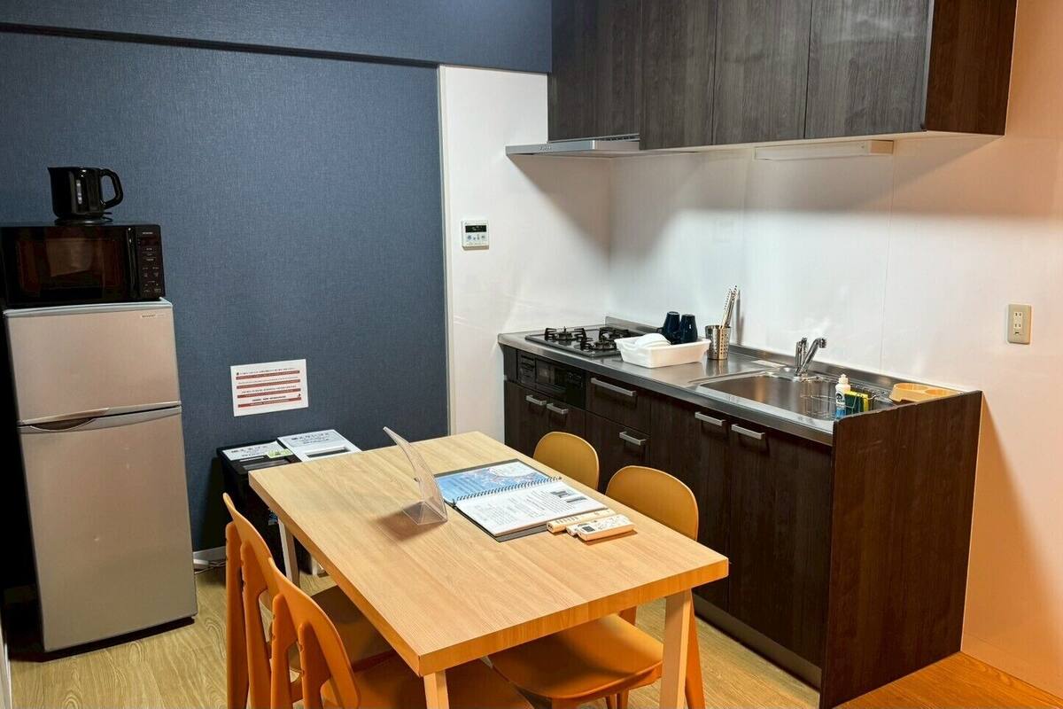 A compact kitchen with a refrigerator, microwave, and sink. There is a wooden table with chairs and a small countertop with a kettle.