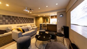 En moderne stue med sofa, skottomaner og sofabord. Der er et køkkenområde med bar og udsigt til vandet.
