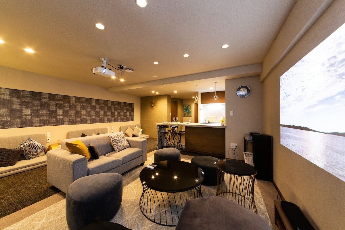 A modern living room with a sofa, ottomans, and a coffee table. There is a kitchen area with a bar and a view of the water.