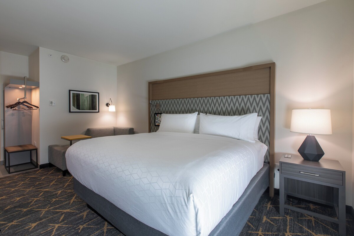 Une chambre d’hôtel avec un grand lit, une table de chevet avec une lampe, une petite table, une chaise, un porte-manteau et un tableau accroché au mur.