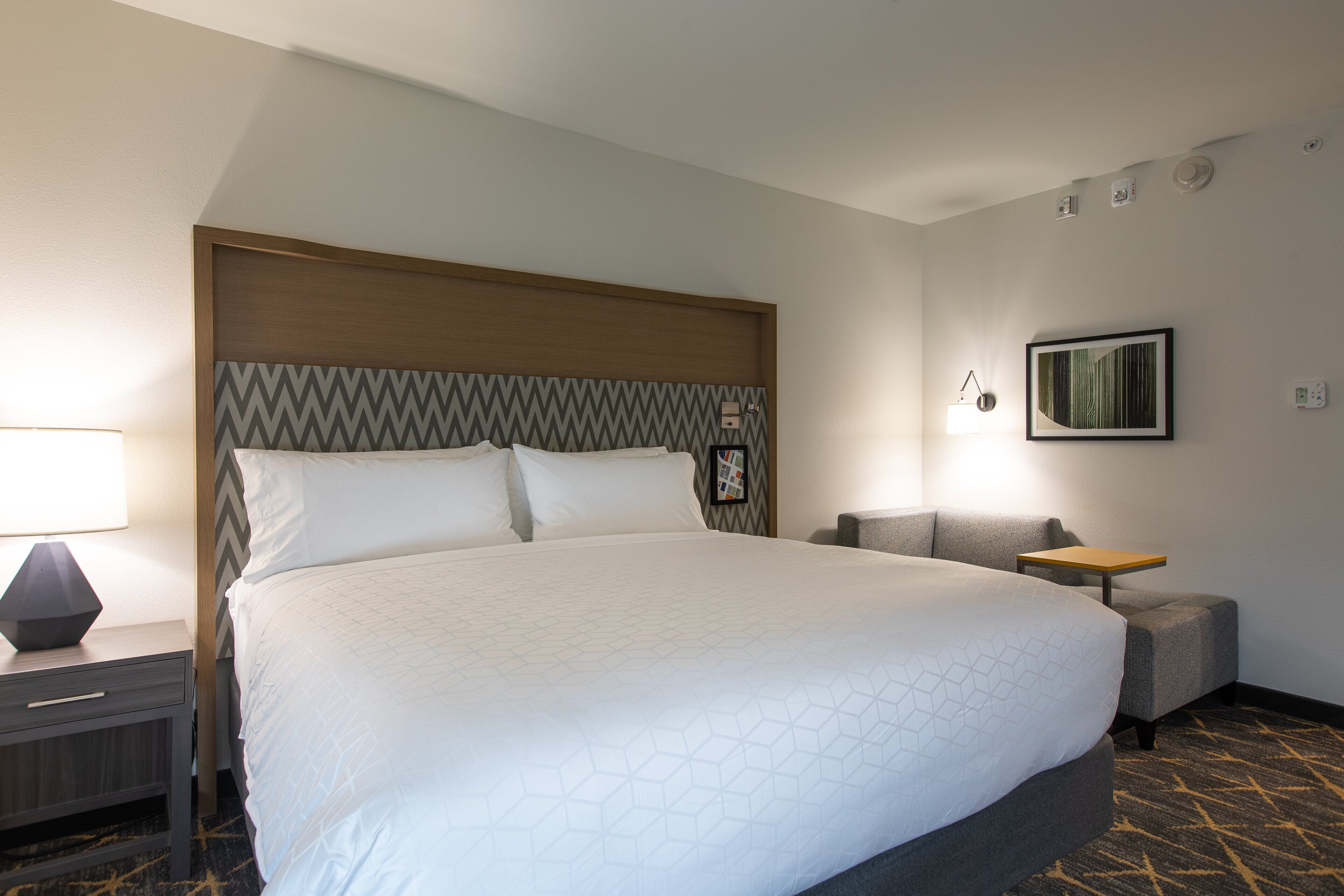A hotel room with a large bed, a nightstand, a lamp, a small table, and a chair.