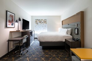 A hotel room with a bed, desk, chair, TV, and a window with a geometric pattern.