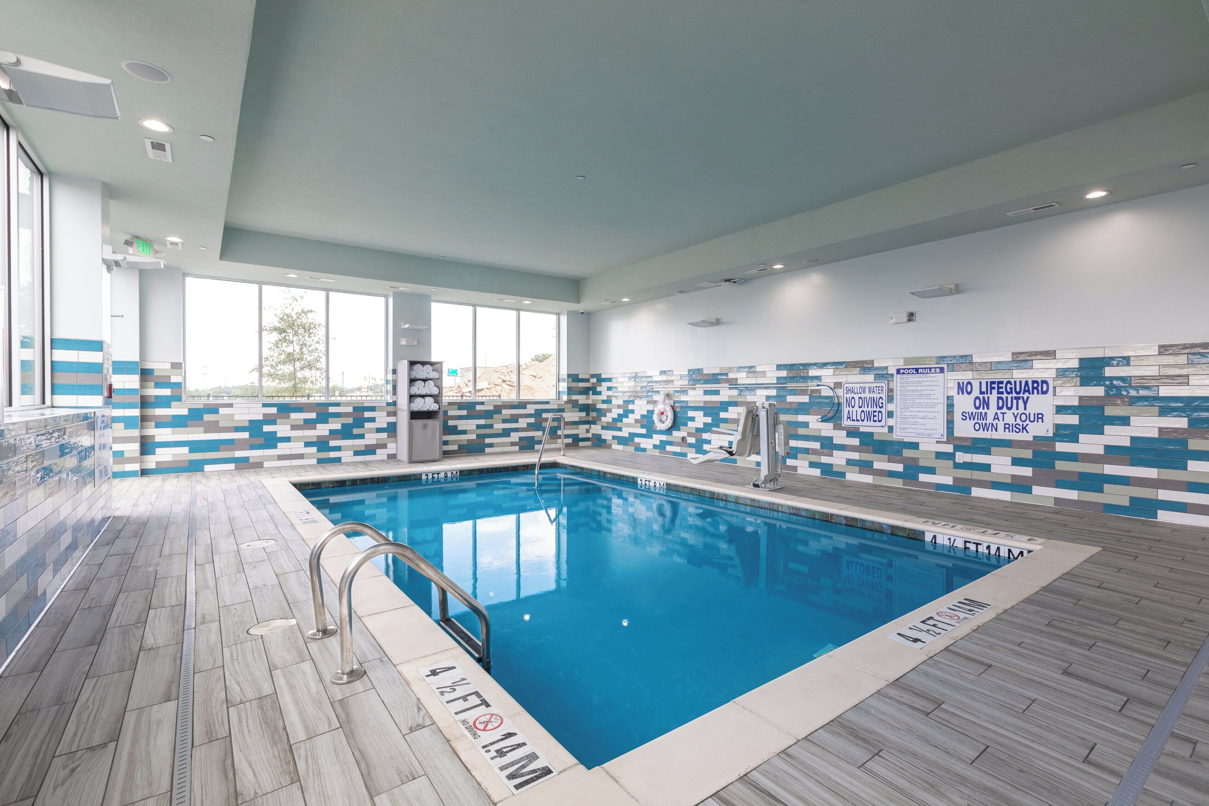 Una piscina interior con normas claras, una silla de socorrista y una pared de azulejos con vistas al exterior.
