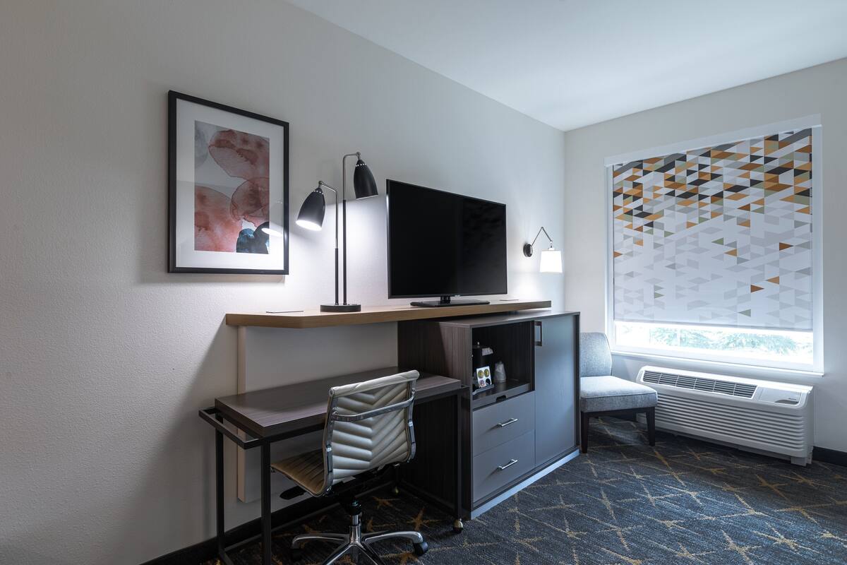 A hotel room with a desk, chair, TV, and a large window with geometric blinds.