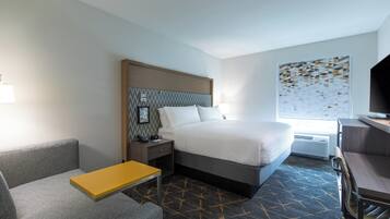 A hotel room with a bed, a desk, a chair, a small table, a nightstand, a lamp, a TV, and a wall art piece.