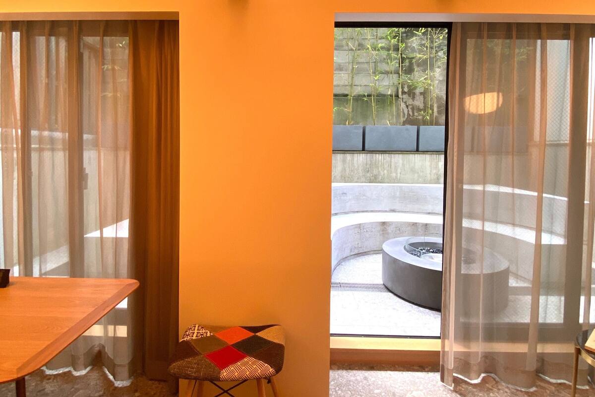 A room with a wooden table, a patterned stool, and a view of an outdoor seating area through a glass door.