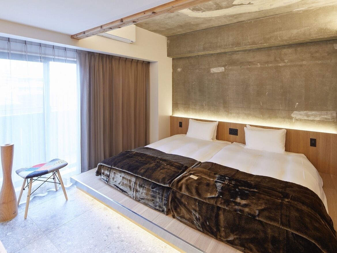 A modern hotel room with a large bed, a wooden headboard, a small stool, and a large window with curtains.