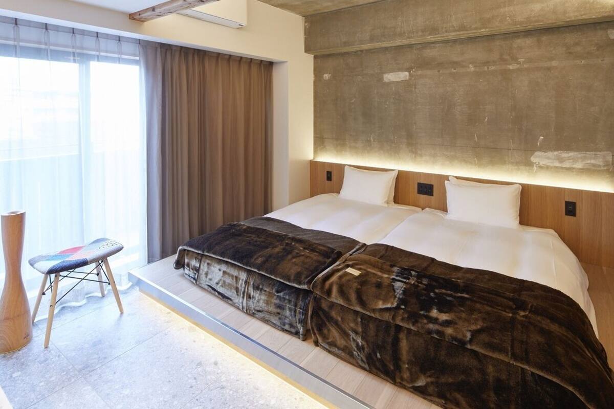 A modern hotel room with a large bed, a wooden headboard, a small stool, and a large window with curtains.