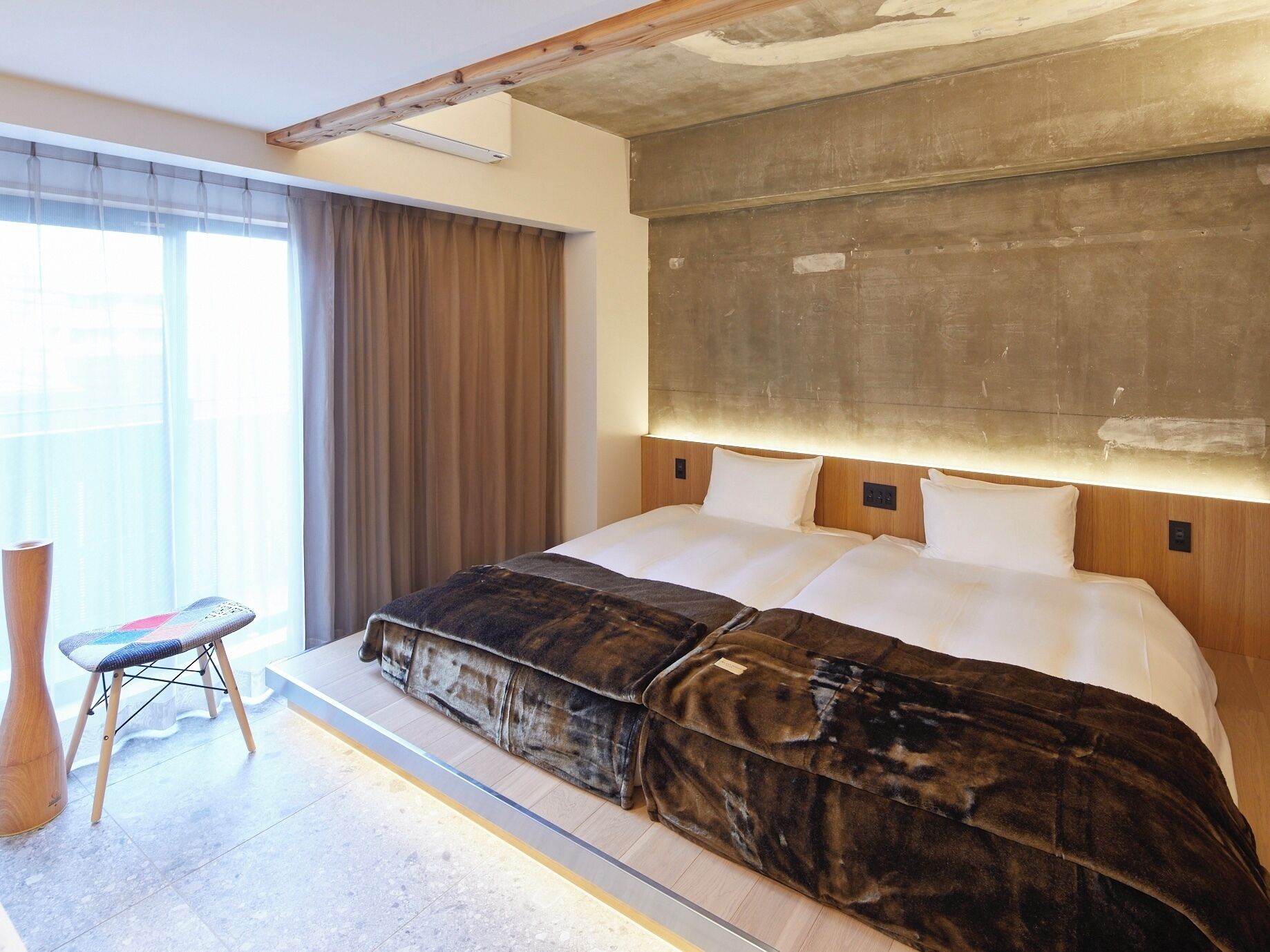 A modern hotel room with a large bed, a wooden headboard, a small stool, and a large window with curtains.