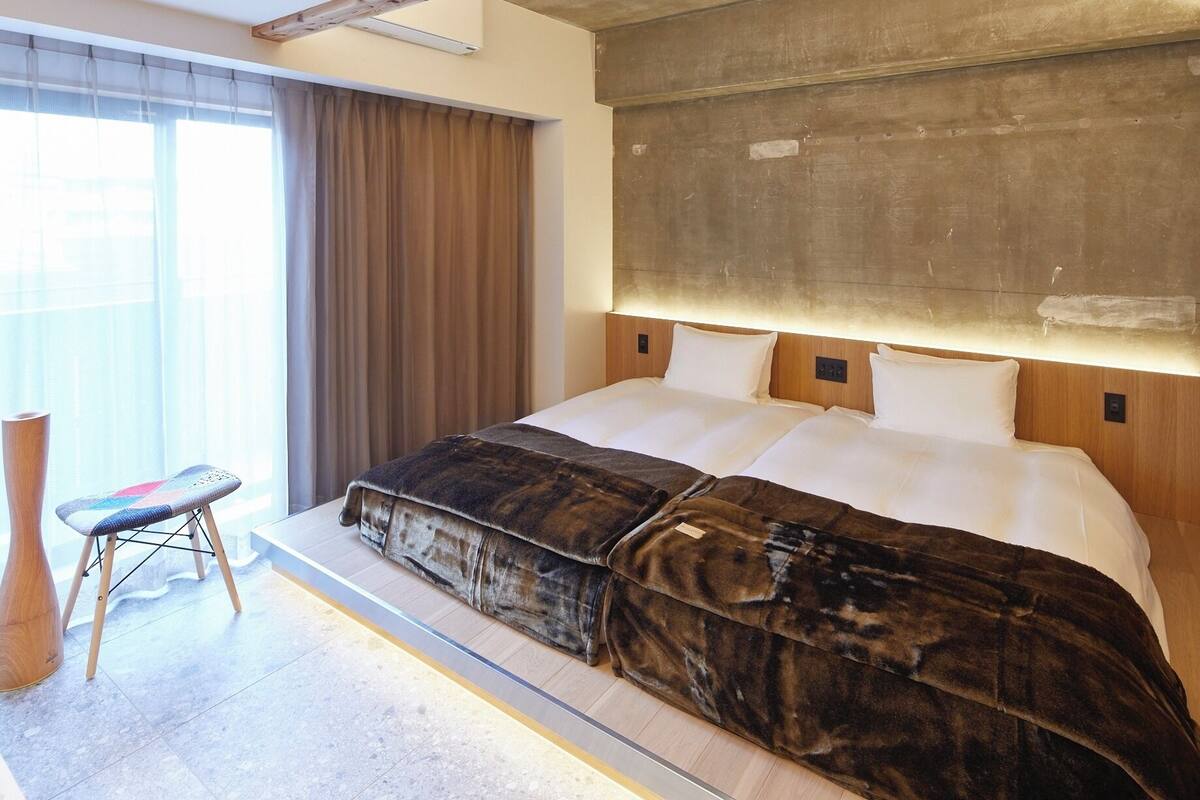 A modern hotel room with a large bed, a wooden headboard, a small stool, and a large window with curtains.