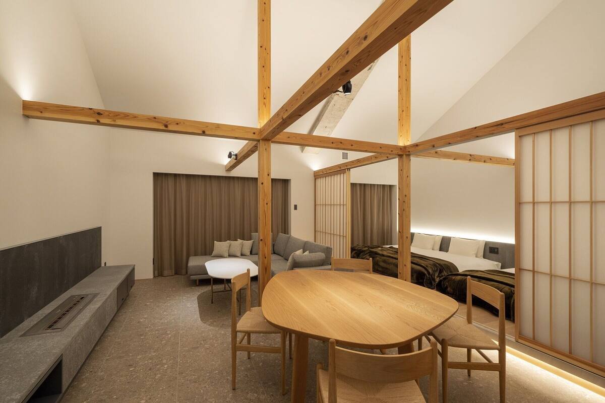 A modern, well-lit room with a wooden ceiling, a round dining table with chairs, a sofa, and a bed with a shoji screen.