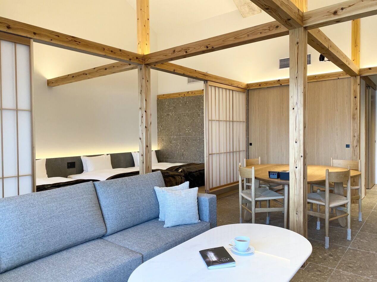 A modern hotel room with a sofa, a dining table with chairs, a bed with a headboard, and wooden beams on the ceiling.