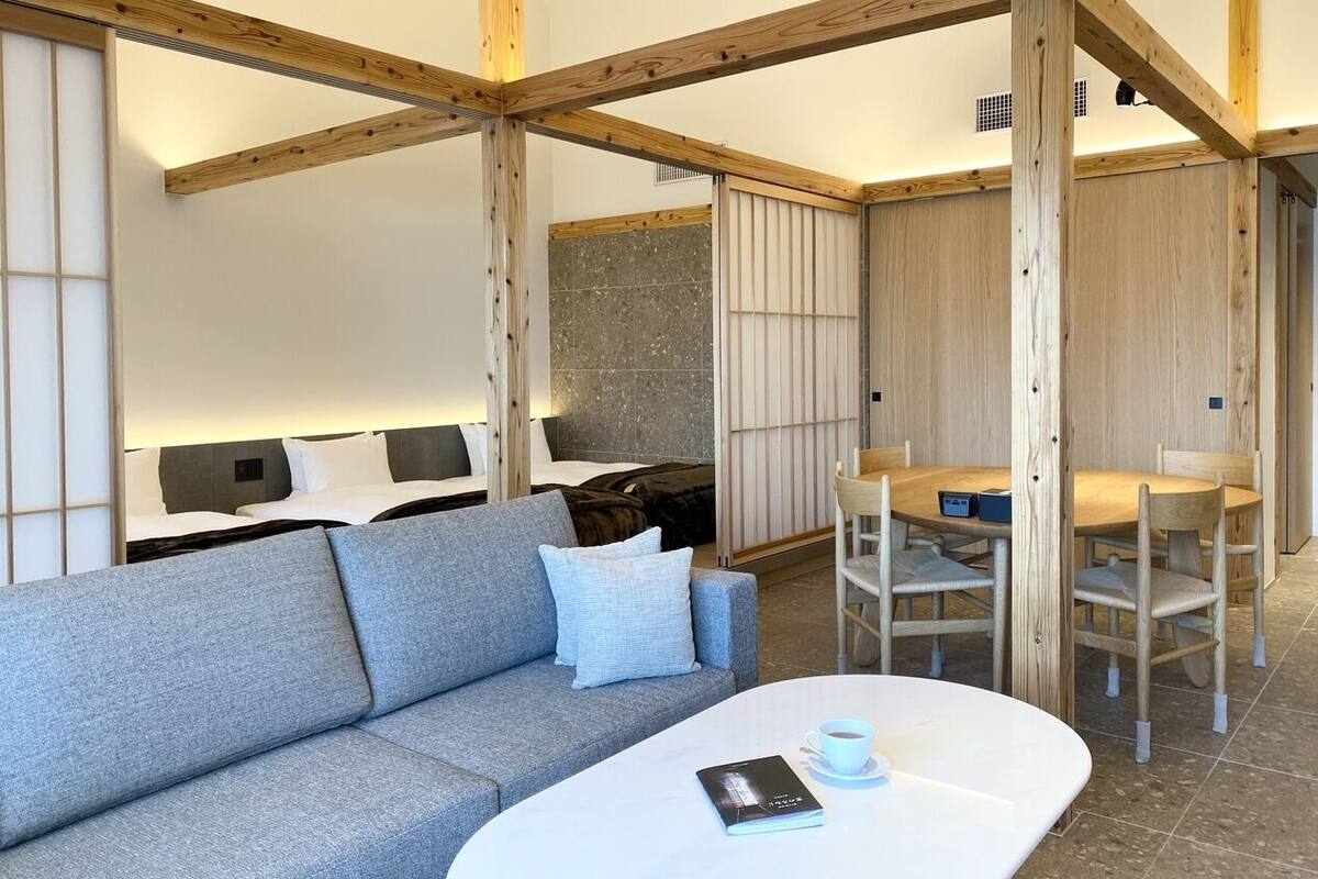 A modern hotel room with a sofa, a dining table with chairs, a bed with a headboard, and wooden beams on the ceiling.
