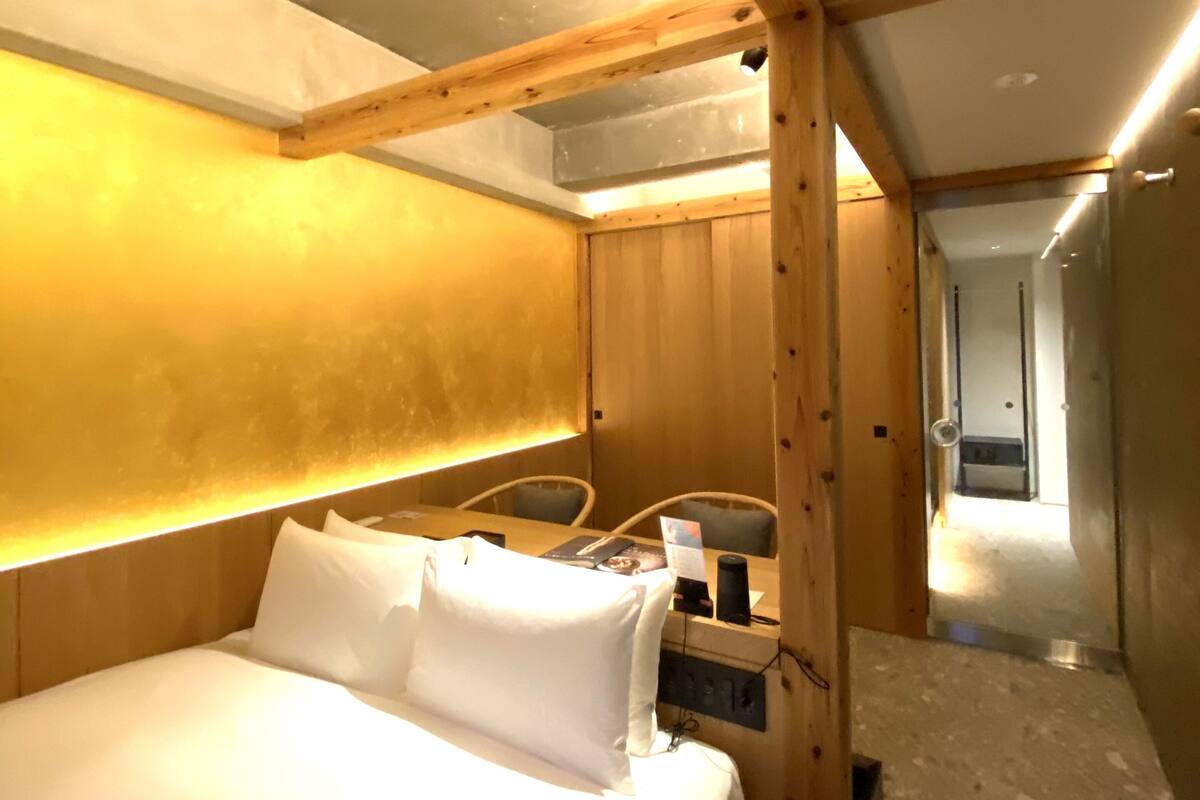 A bedroom with a large bed, wooden beams, a headboard with built-in lighting, and a view of a bathroom through an open door.