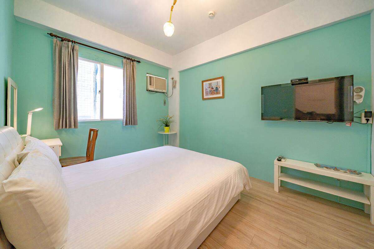 Une chambre avec un grand lit, une télévision, une table de chevet, une chaise, une fenêtre avec des rideaux et un appareil de climatisation.