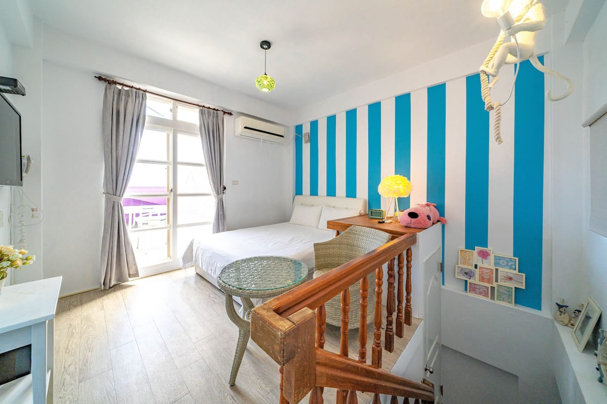Une chambre à coucher avec un lit, une table en verre, une chaise en osier, une chaise en bois, un escalier et un mur à rayures bleues et blanches.