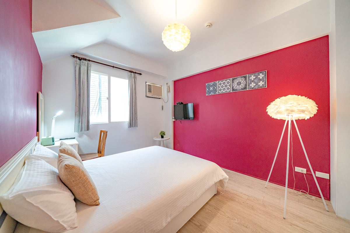 Une chambre avec un grand lit, un mur d’accent rouge, un luminaire au plafond et une lampe sur pied.