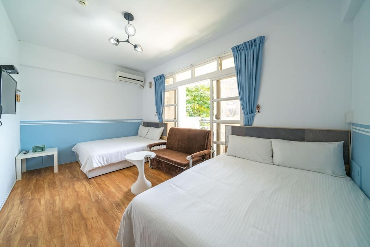 Une chambre d’hôtel avec deux lits, un coin salon comprenant un fauteuil, une petite table et un ventilateur de plafond.
