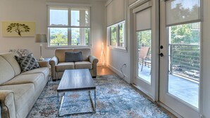 Un salón amplio con un sofá, una mesa de centro y una lámpara. Hay ventanas grandes y una puerta corrediza que da a un área exterior.