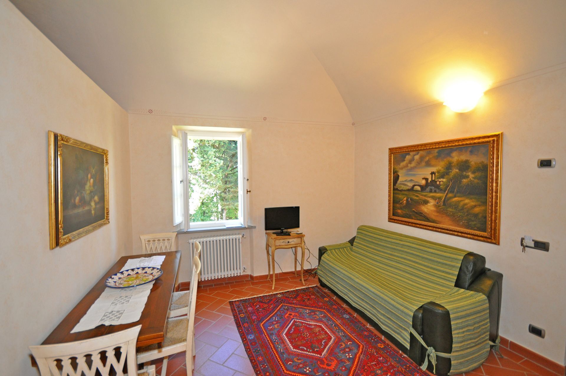 A room with a wooden table, a green sofa, a red patterned rug, a window with a view of trees, and two paintings on the walls.