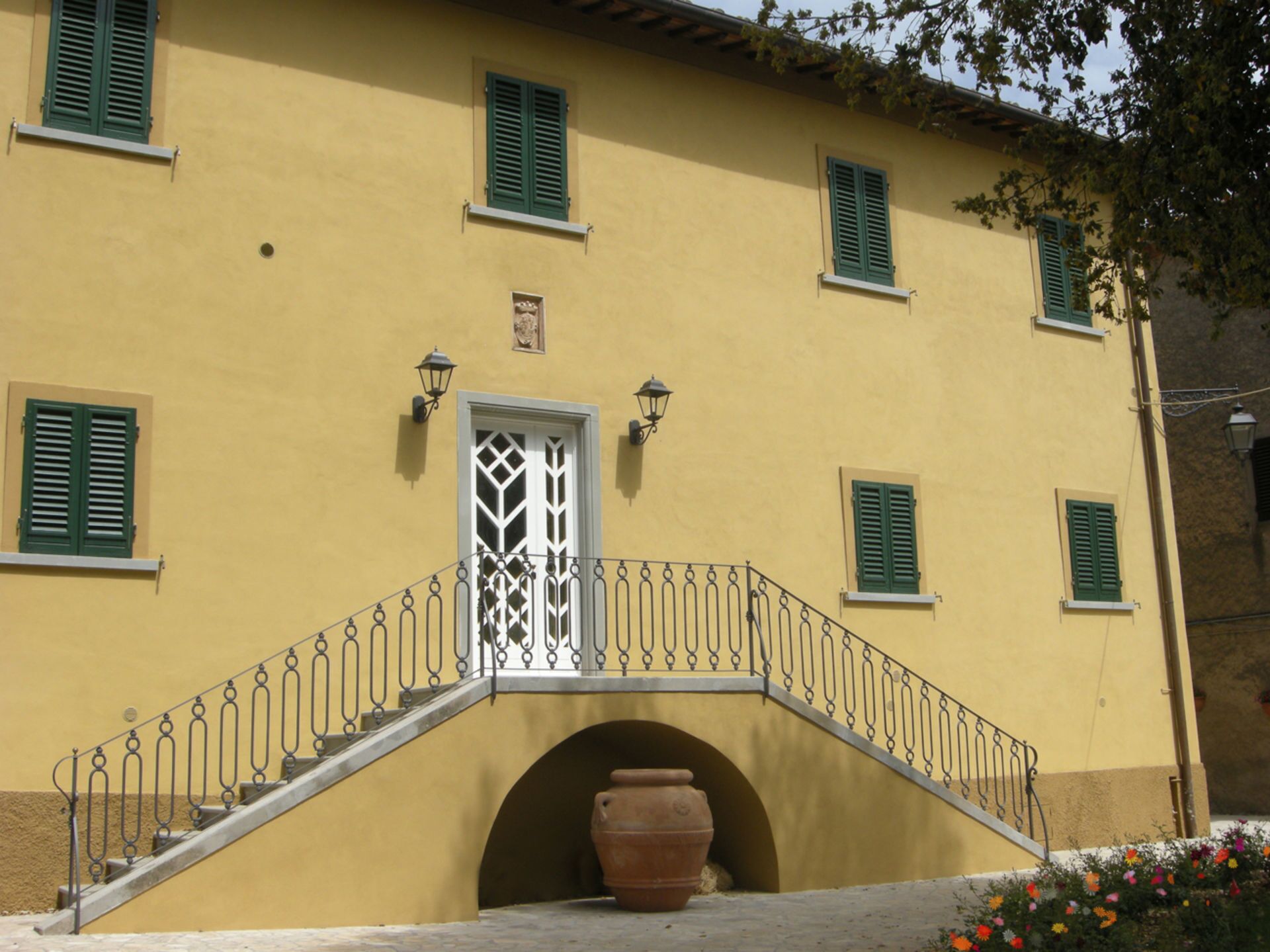Ein gelbes Gebäude mit grünen Fensterläden, einer weißen Tür mit dekorativem Muster und einem kleinen Balkon mit Metallgeländer.