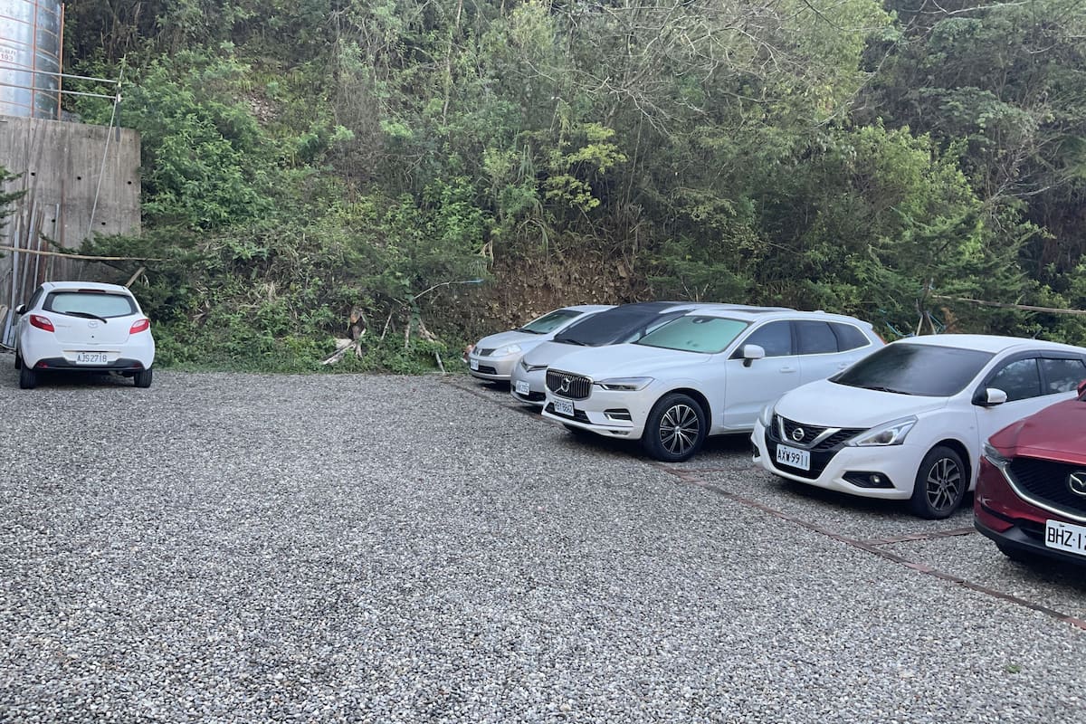A parking area with several cars, a gravel surface, and a forested hillside in the background.