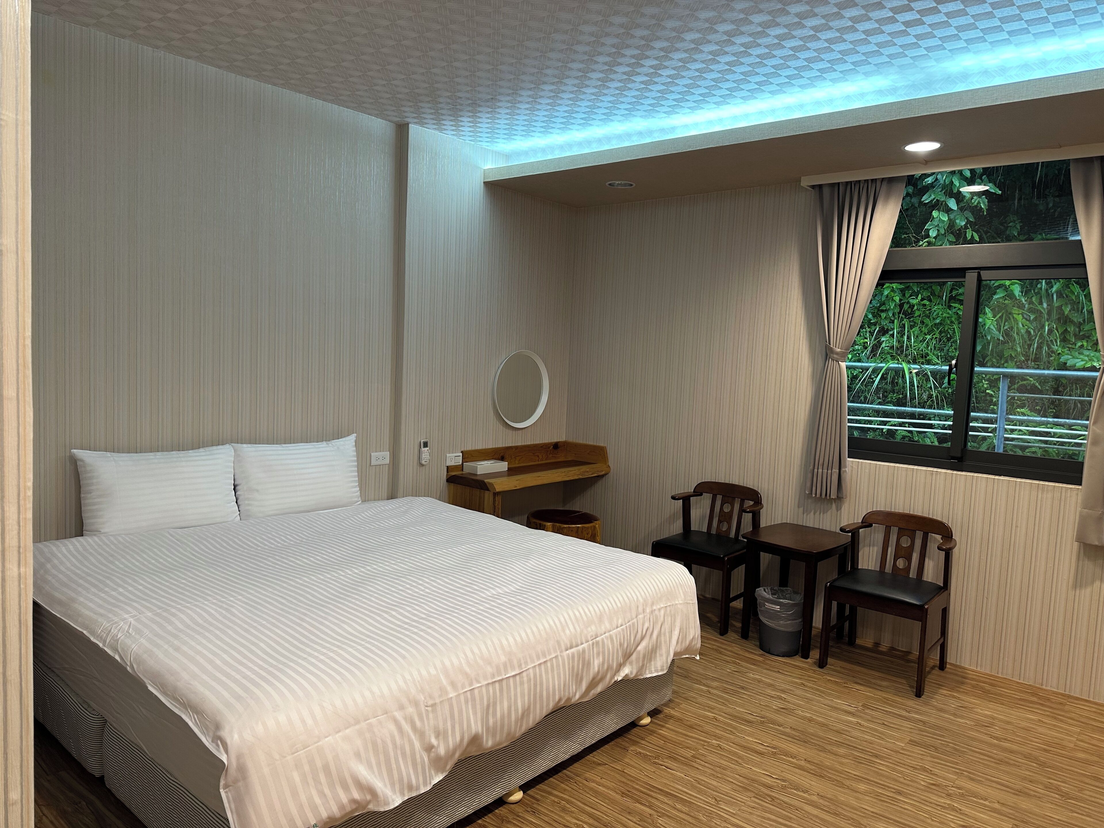 A hotel room with a large bed, a wooden desk, two chairs, a small table, a mirror, and a window with a view of greenery.