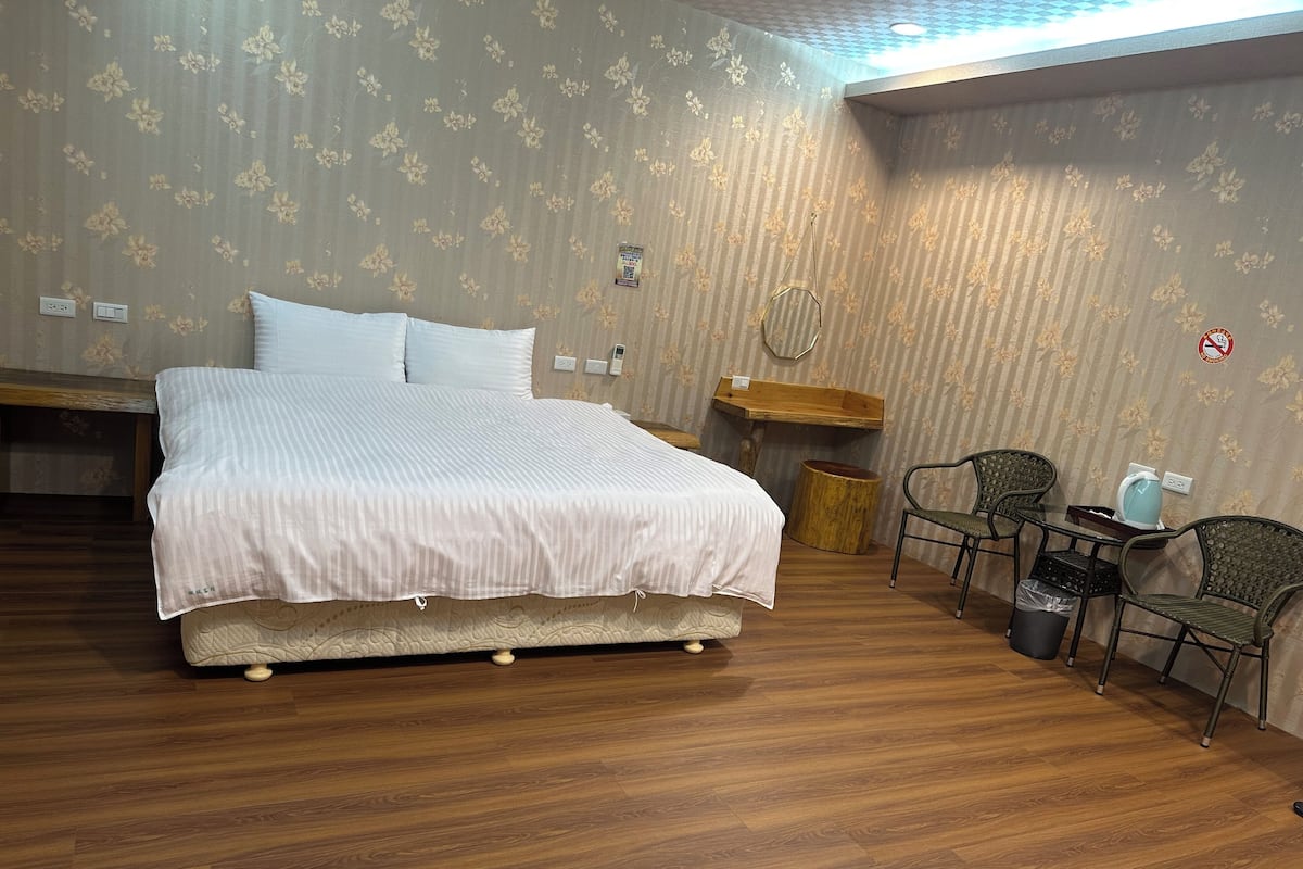A bedroom with a bed, a wooden nightstand, a chair, a small table, and a trash bin.