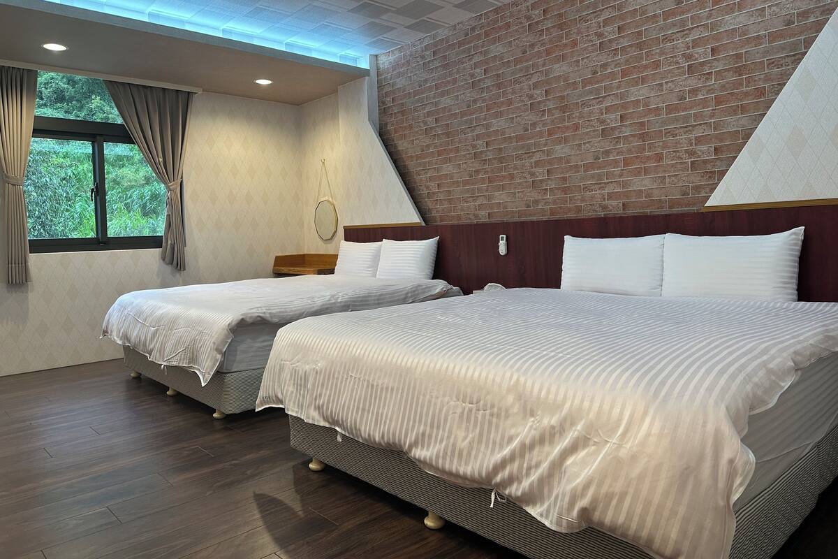 A hotel room with two beds, a brick wall, a window with curtains, and a ceiling with recessed lighting.