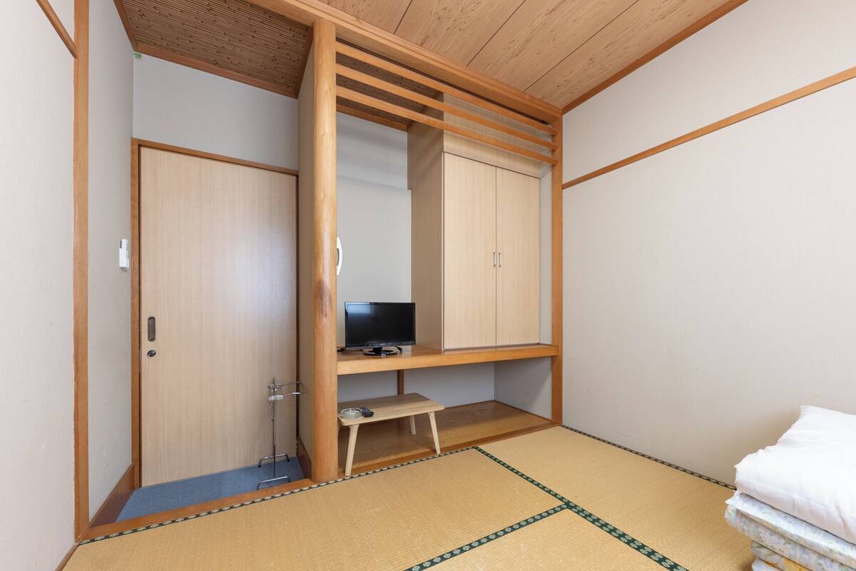 Une pièce avec une armoire, un téléviseur posé sur une étagère, un lit avec du linge de lit blanc et un sol recouvert de tatamis.