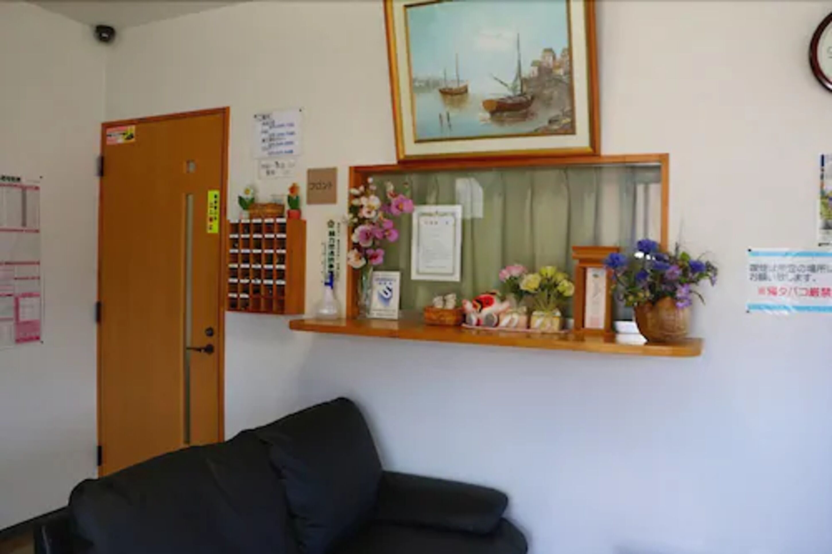 A room with a wooden door, a black sofa, a shelf with flowers and a framed picture, and a bulletin board with notices.