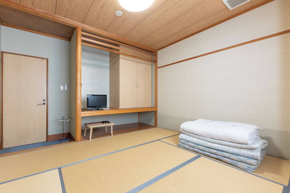 Une pièce de style japonais traditionnel, avec un sol recouvert de tatamis, une télévision, une armoire et un lit dont la literie est soigneusement pliée.