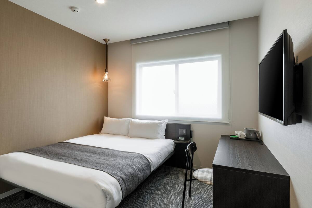 A hotel room with a bed, a desk, a chair, a TV, and a window.