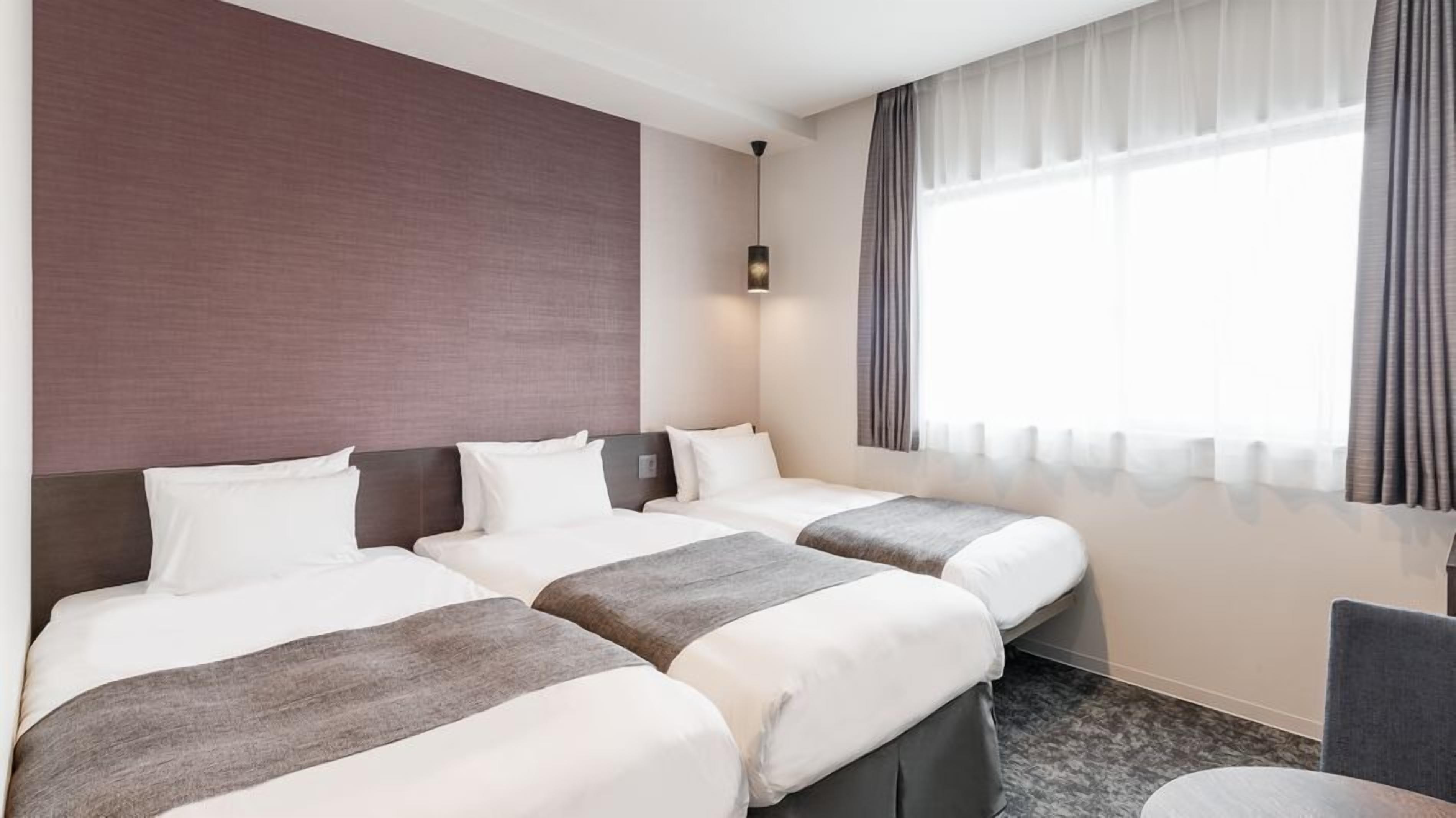 A hotel room with two beds, a large window with curtains, a wall-mounted light, and a chair.