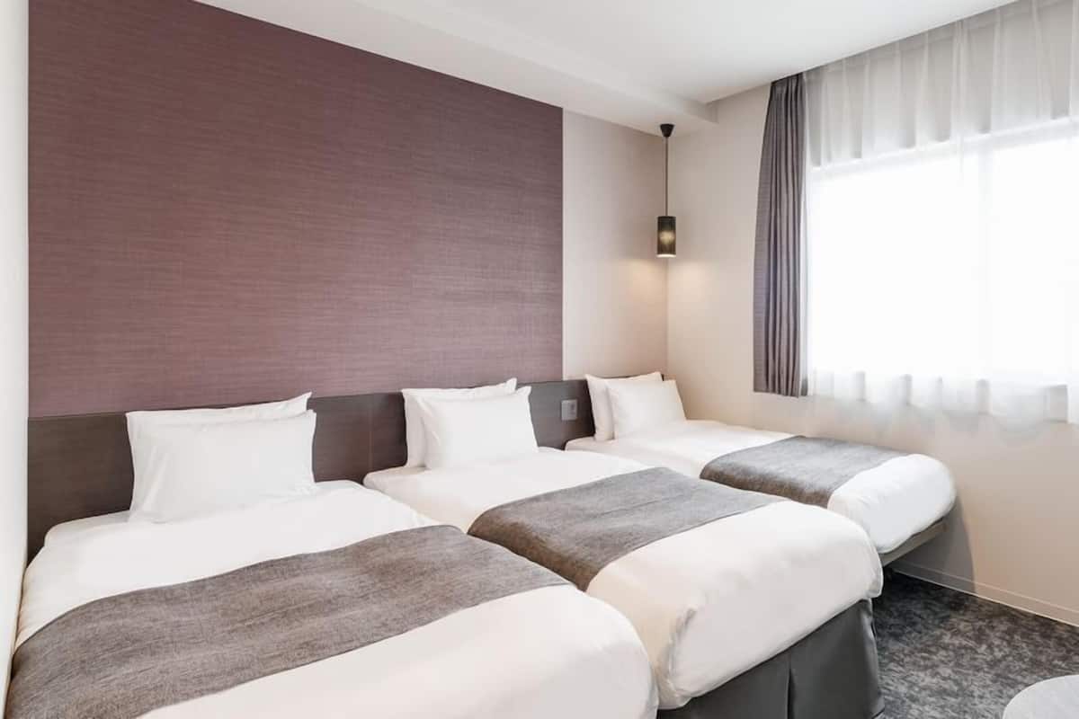 A hotel room with two beds, a large window with curtains, a wall-mounted light, and a chair.
