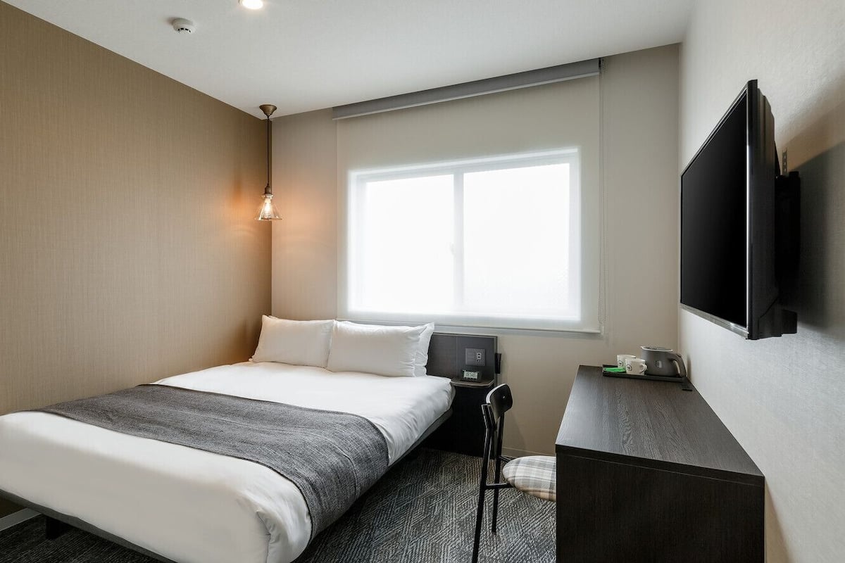 A hotel room with a bed, a desk, a chair, a TV, and a window.