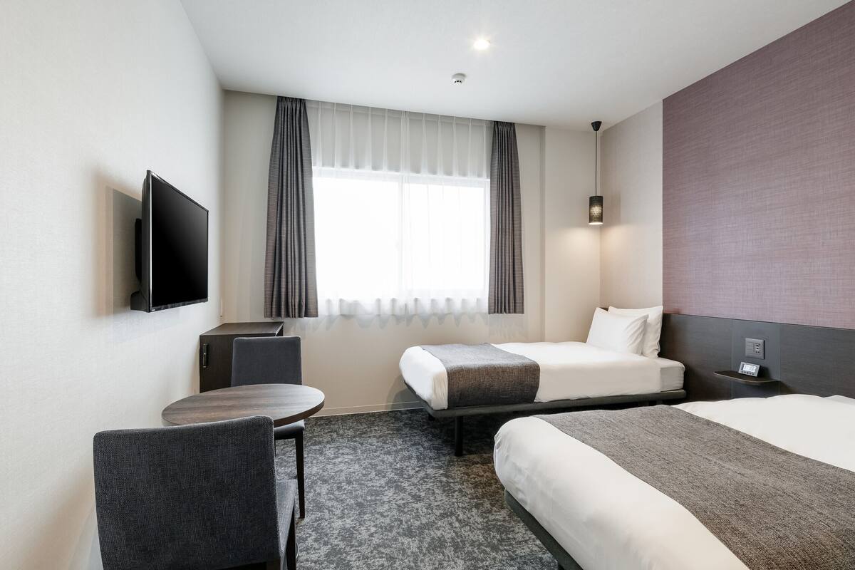 A hotel room with two beds, a desk, a TV, and a window with curtains.