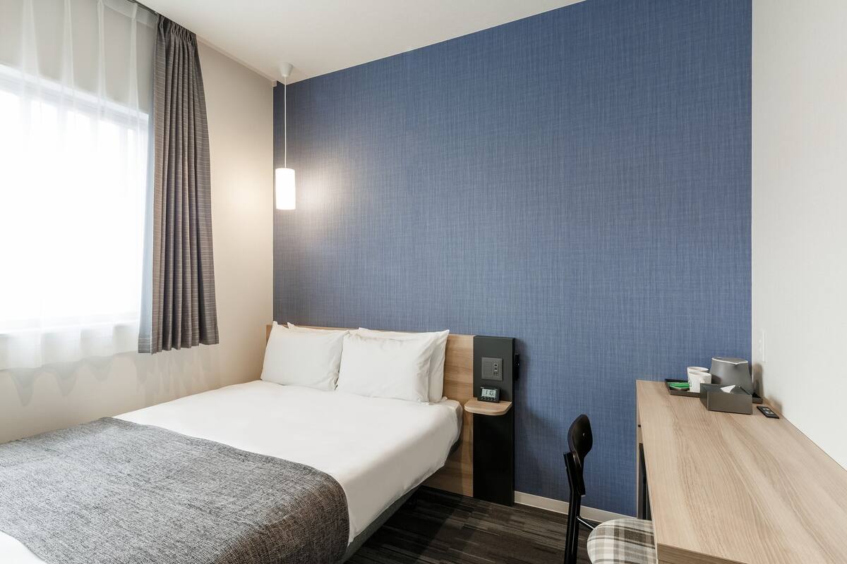 A hotel room with a bed, a desk, a chair, a nightstand, and a wall-mounted control panel.