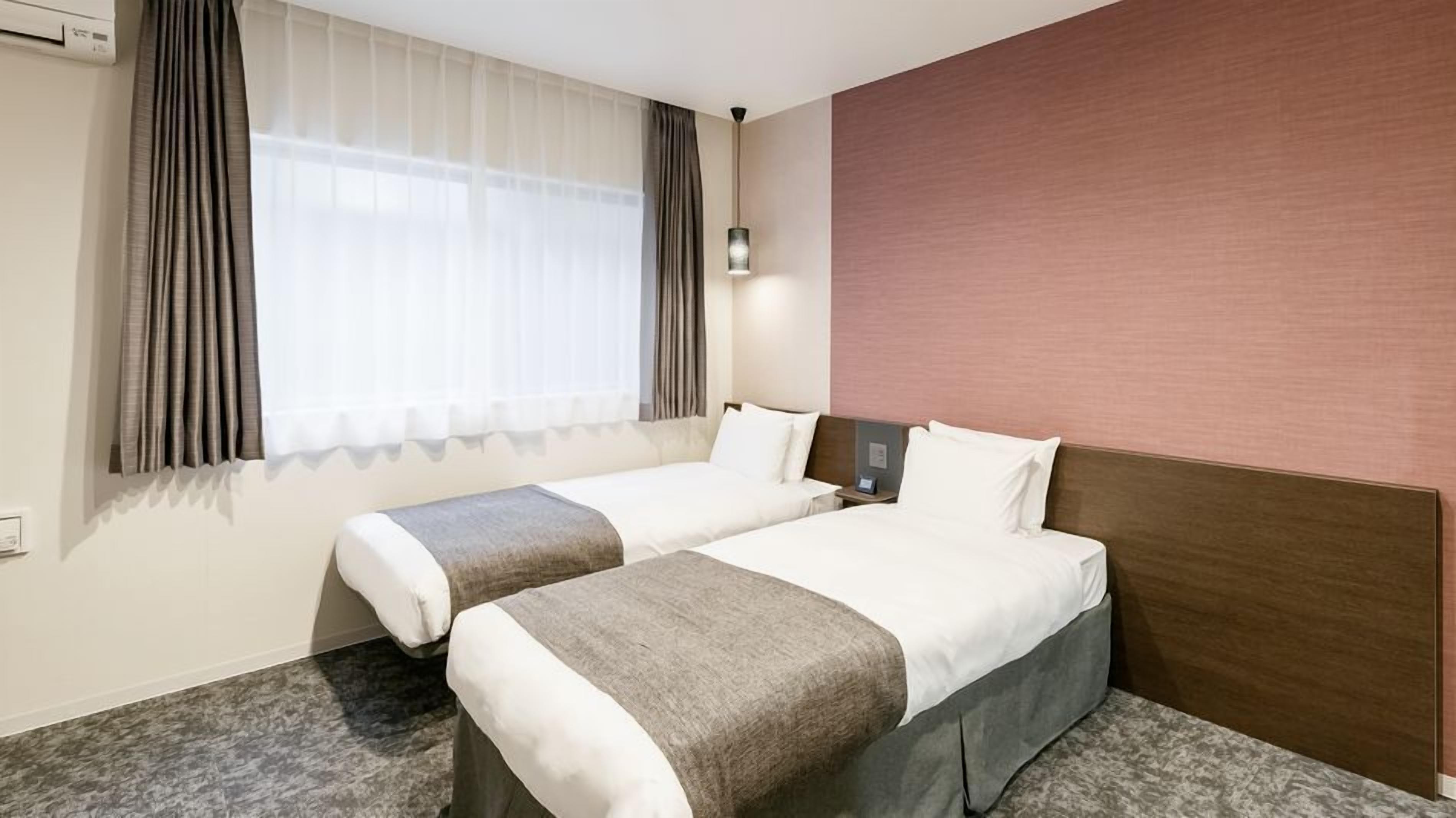 A hotel room with two beds, a large window with curtains, a wall-mounted lamp, and a carpeted floor.