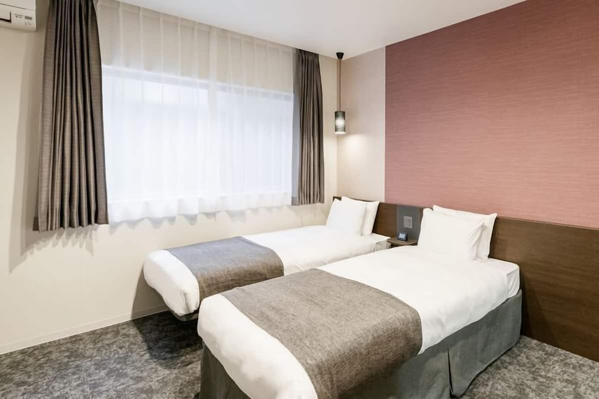 A hotel room with two beds, a large window with curtains, a wall-mounted lamp, and a carpeted floor.