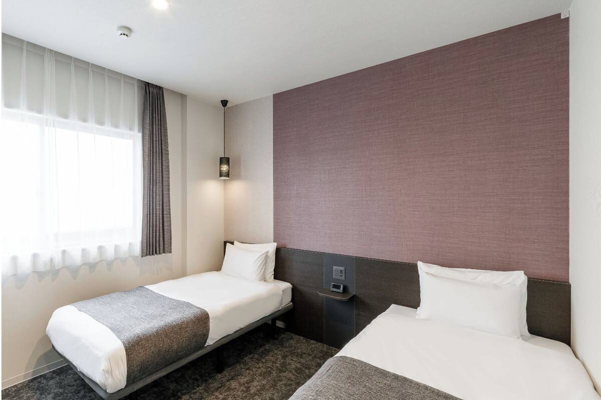 A hotel room with two beds, a bedside table, a lamp, and a window with curtains.