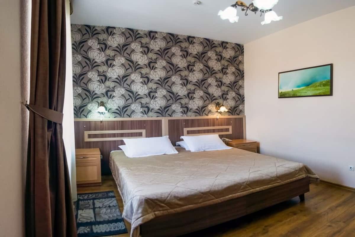 A bedroom with a large bed, wooden headboard, two bedside tables, a nightstand, a patterned wallpaper, a painting on the wall, and a wooden floor.