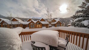 Cubierta nevada con una mesa y sillas, con vista a una hilera de cabañas de madera con tejados cubiertos de nieve.