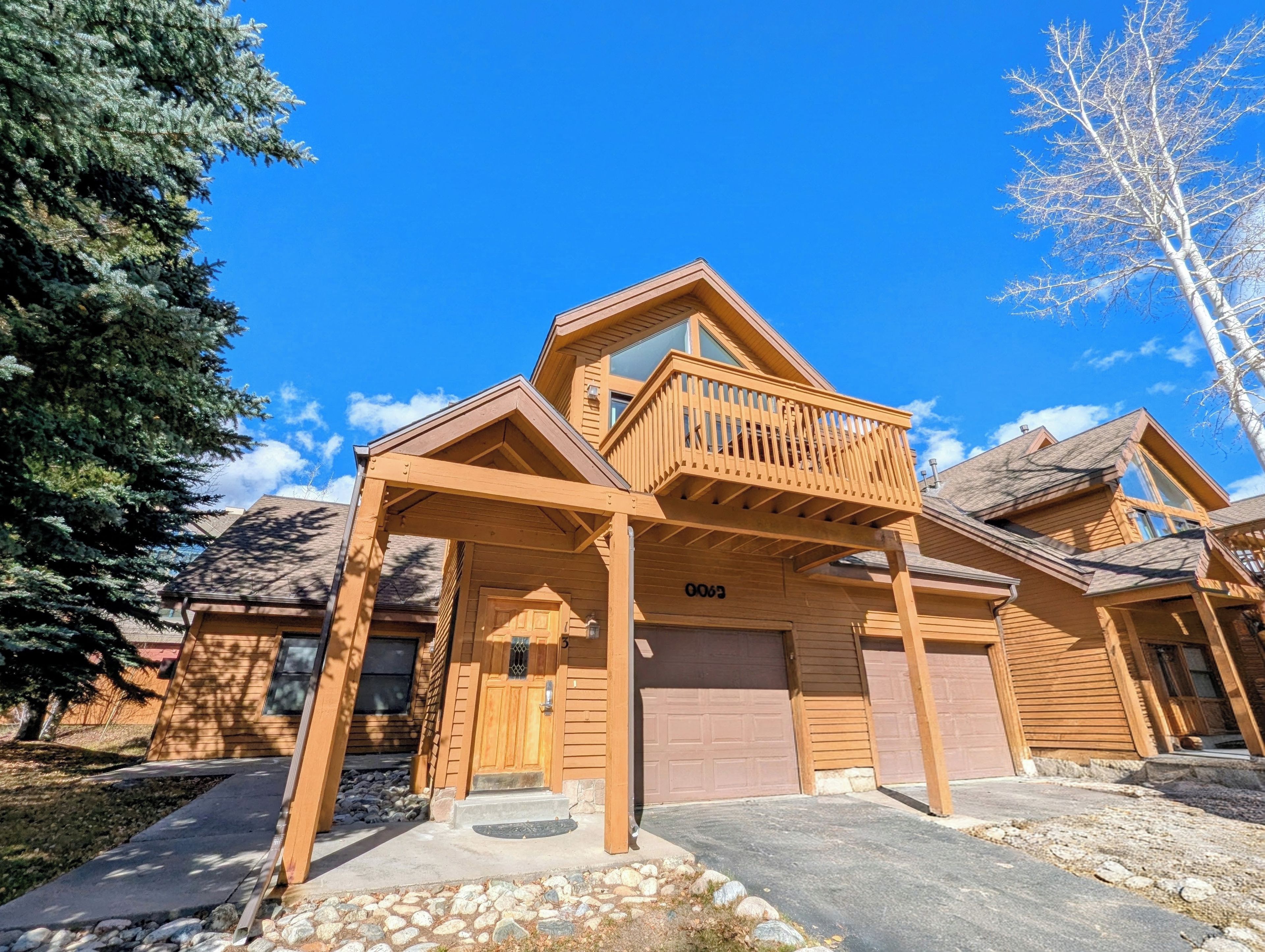 This charming wood-sided cabin stands ready in the crisp mountain air, its warm timber façade and covered balcony framed by towering evergreens and a brilliant blue sky.